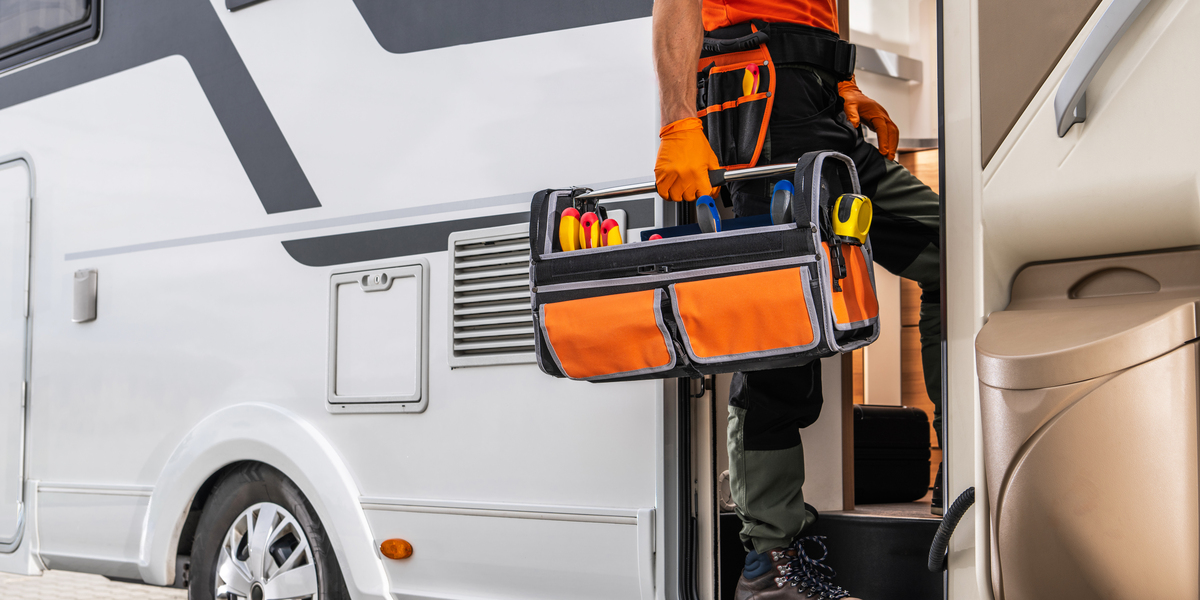 A technician wearing an orange shirt and work pants is entering a motorhome while carrying a toolbox. The toolbox is full of tools, including screwdrivers and other items.